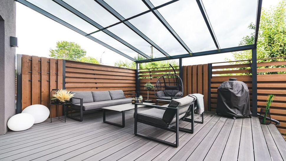 Covered patio with gray lounge seating, black grill, wood deck, and wooden privacy fence.