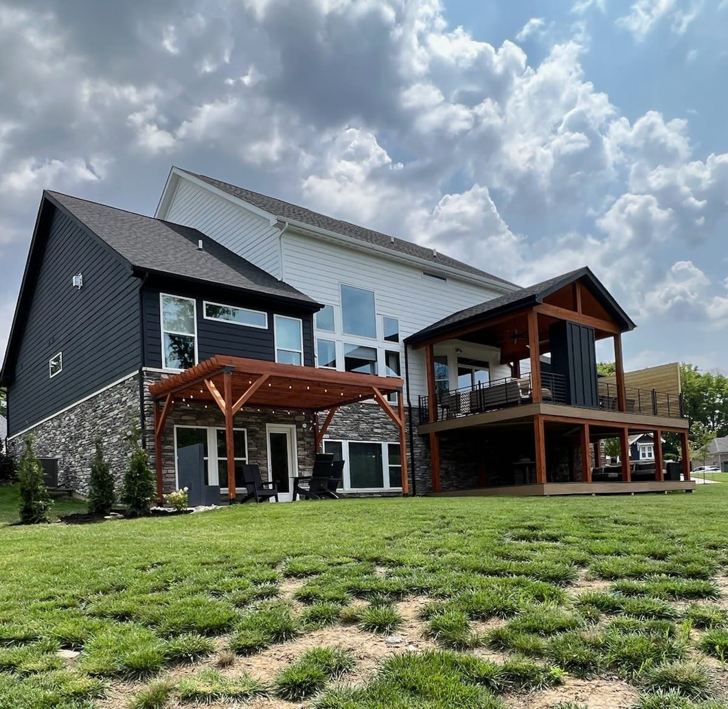 Modern house exterior with dark siding, wood decks, and large windows on a grassy hill under a cloudy sky