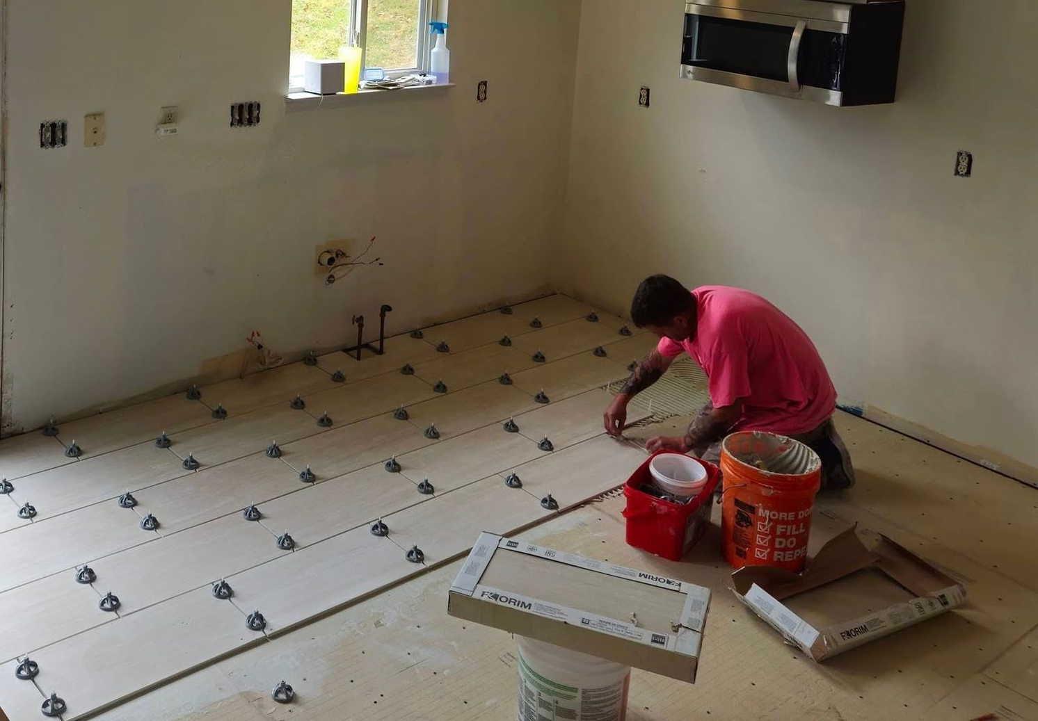 Worker tiling a room floor with white square tiles, buckets and tools nearby