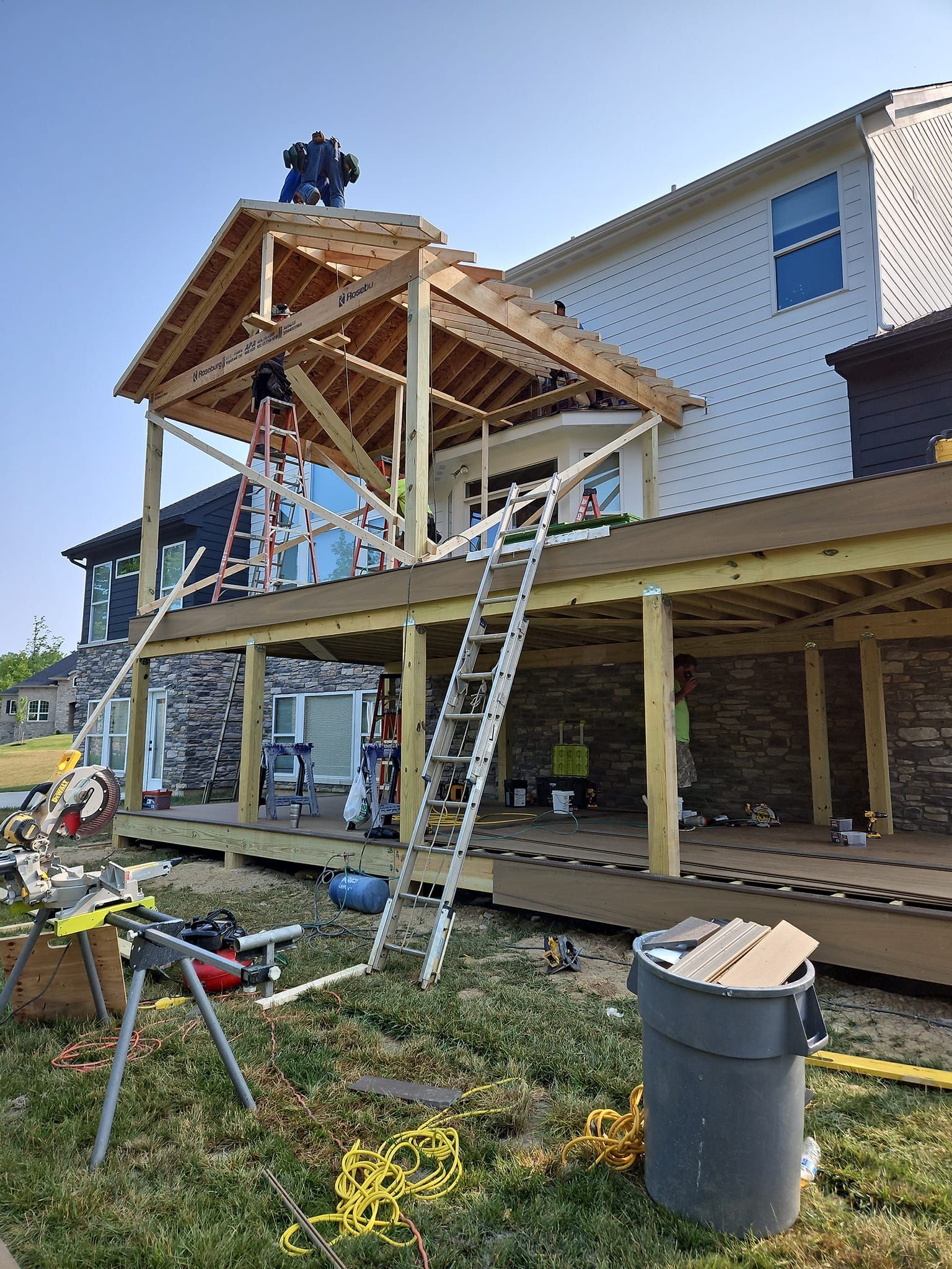 People building a wooden porch and roof frame on a house, with ladders and construction tools.