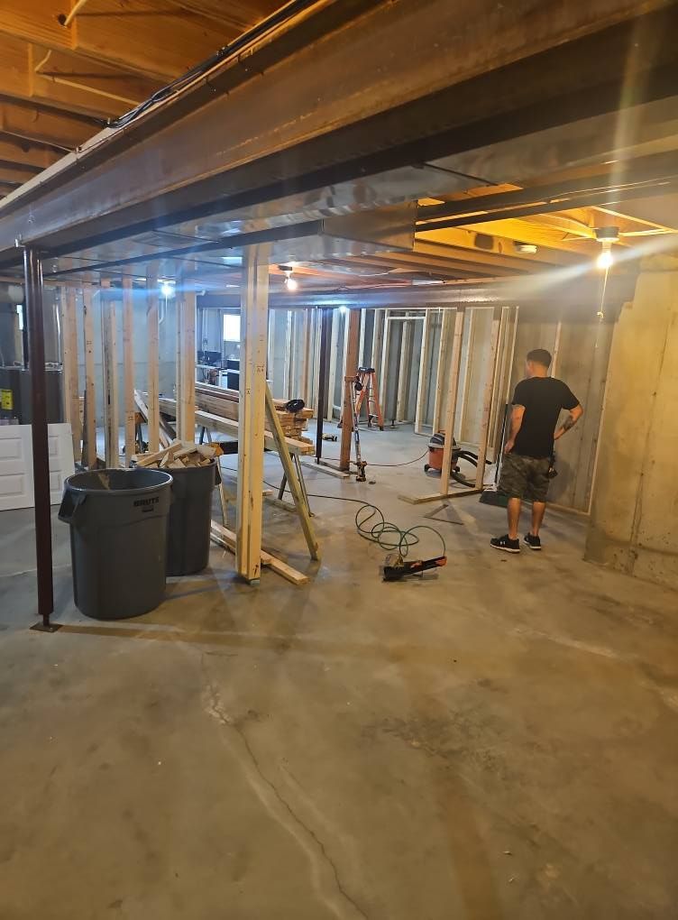 Basement under construction with exposed beams, temporary supports, tools, and two workers floor painting