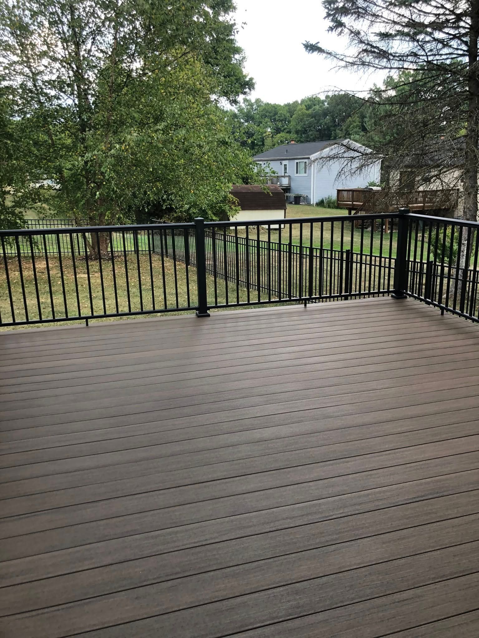 Wooden deck overlooking a yard with trees, a black railing, and houses in the distance