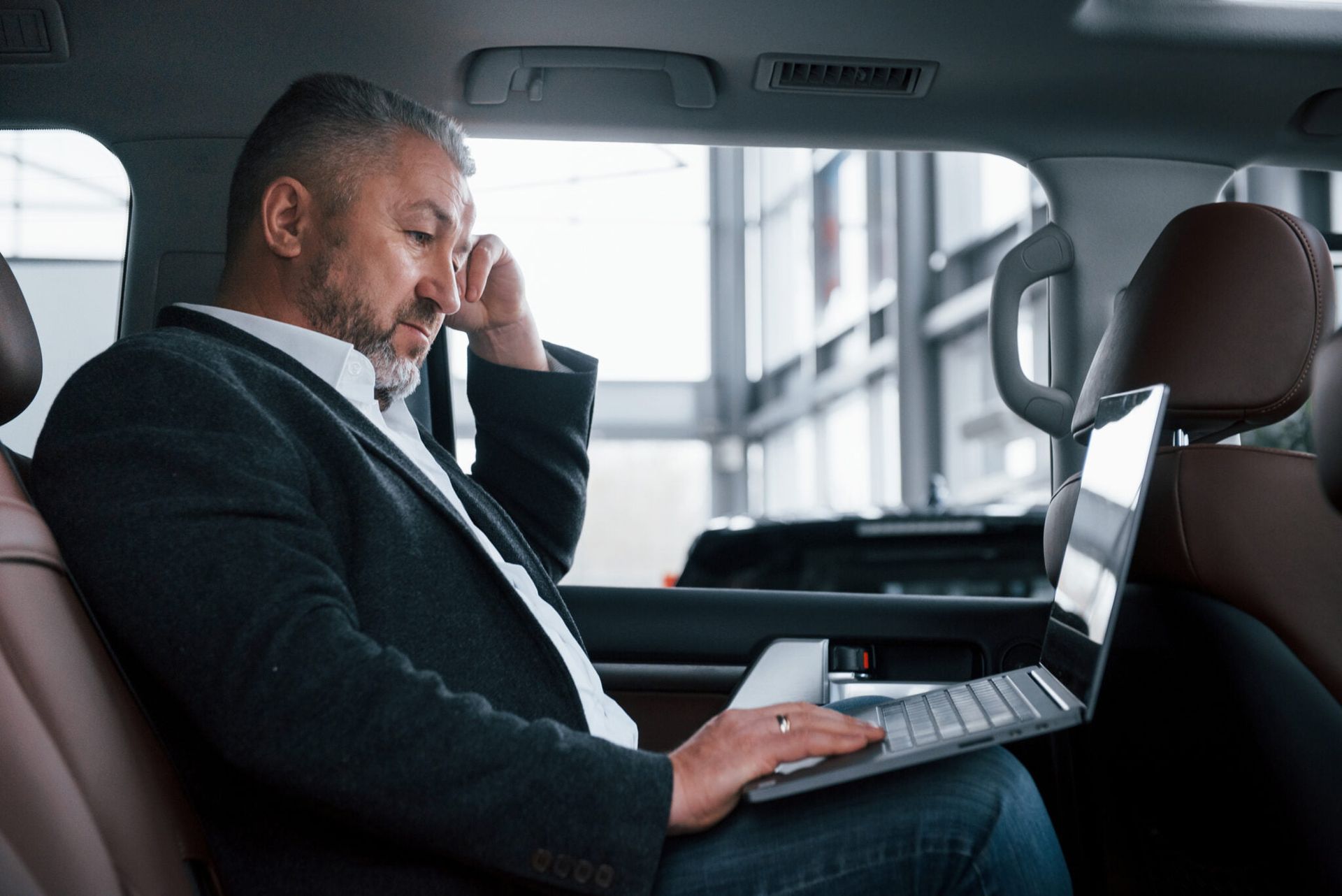 A man is sitting in the back seat of a car using a laptop and talking on a cell phone.