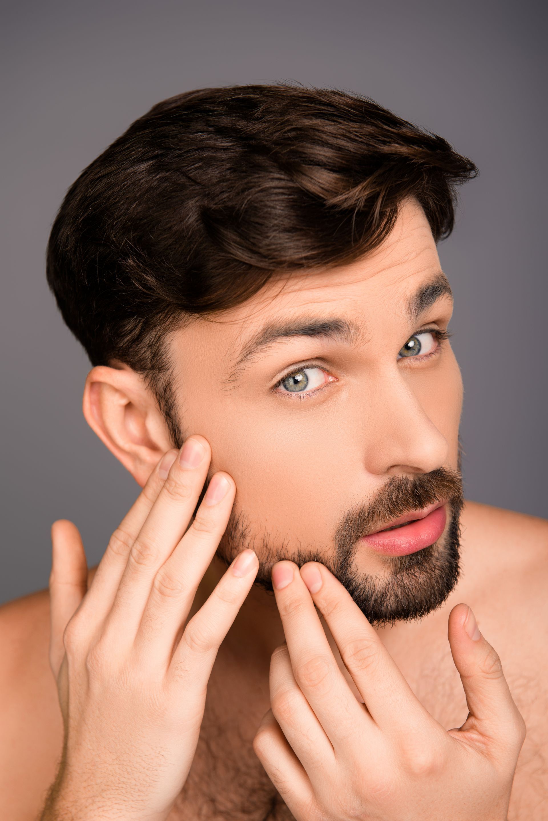A shirtless man with a beard is looking at his face.