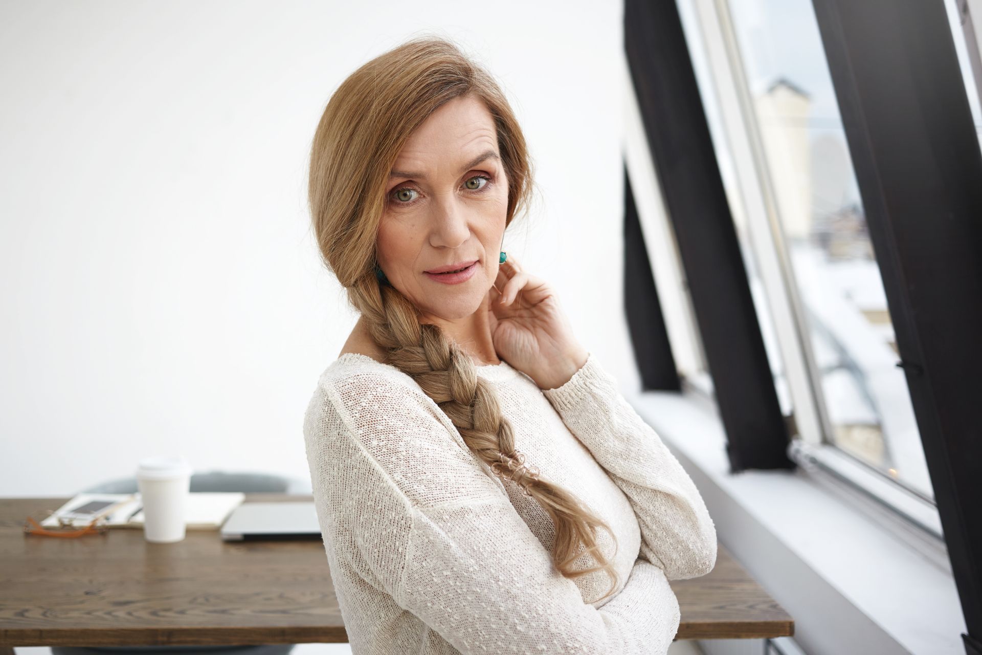 A woman in a white sweater is standing in front of a window.