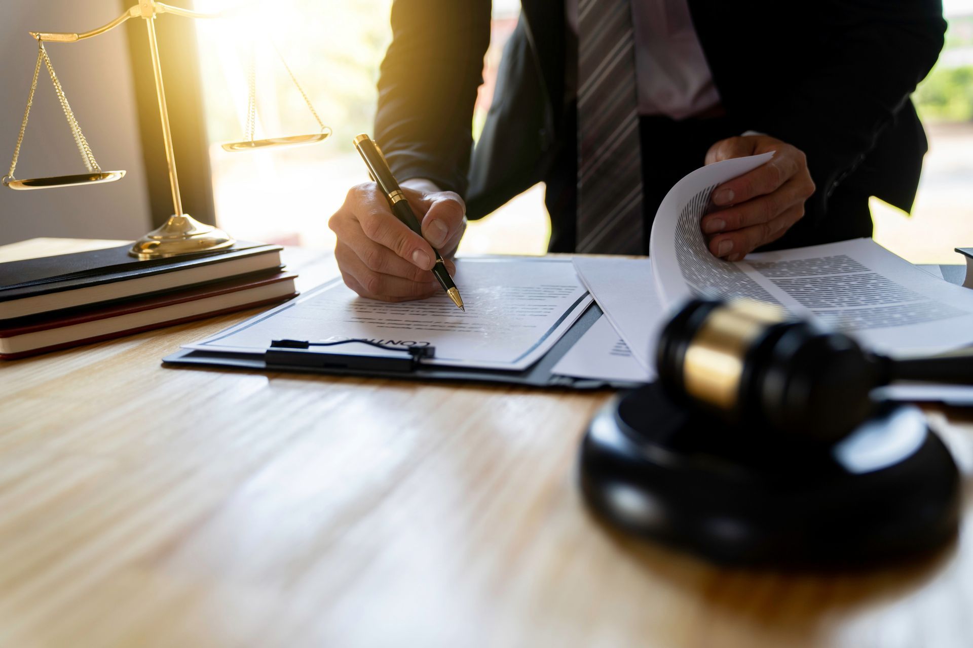 A lawyer in a suit signing a document on a desk, gavel and scales of justice nearby.