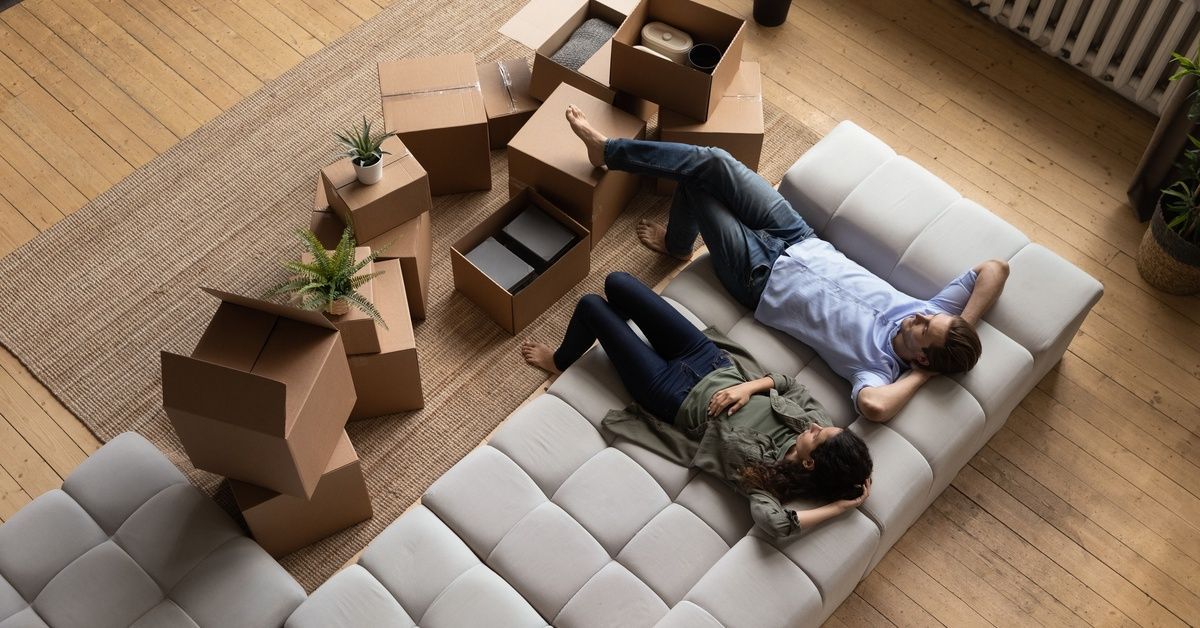 A man and a woman are sitting back on a large couch and relaxing. There are boxes strewn all around