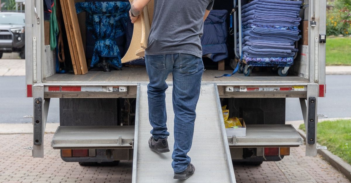 A man in a dark shirt is lifting a table into a moving truck. The man is walking up a ramp.