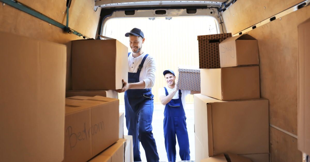 Two men, both of whom are wearing overalls, are lifting boxes and putting them into a van.