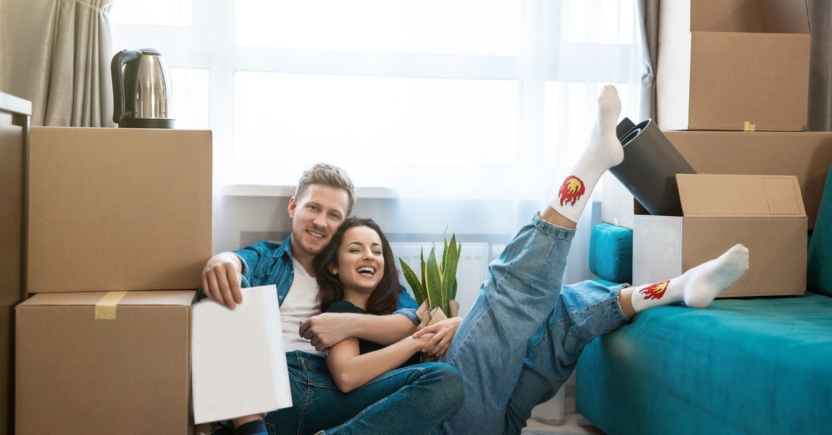 A man and a woman are sitting on a floor, cardboard boxes stacked high surround them. They are both