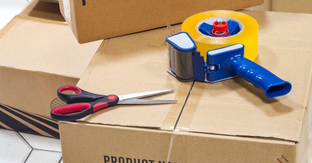 A tape dispenser is sitting on top of a cardboard box. There is a pair of scissors next to the tape dispenser.