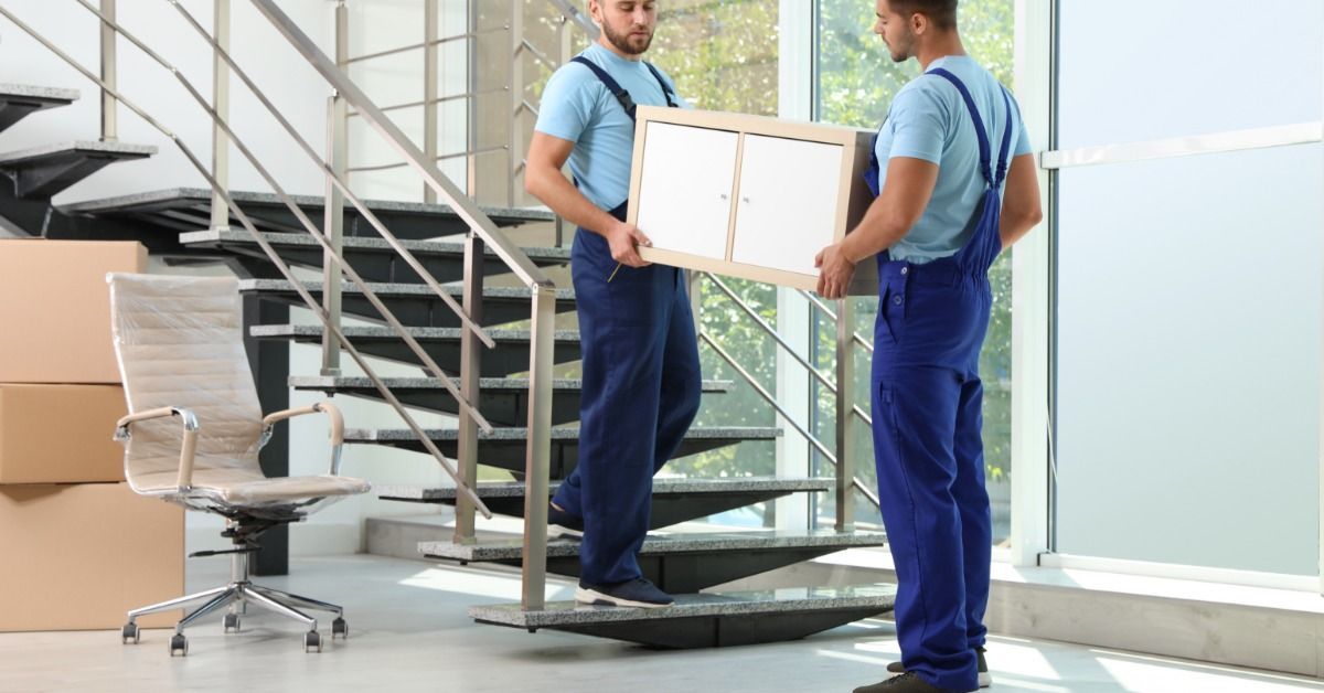Two men in dark blue overalls and light blue shirts are carrying a box down a flight of stairs.