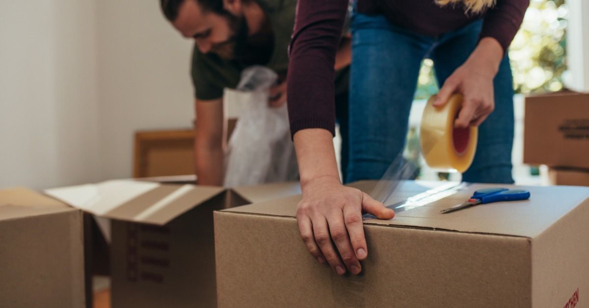 A woman is taping a box while a man behind her is kneeling down and packing items into a separate box.