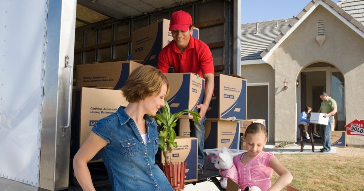 A man in a red uniform is unloading boxes from the back of a moving truck. There is a woman and children by the truck.