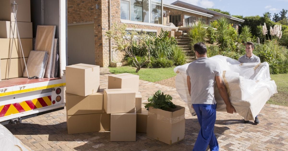 Two men are lifting a couch together. One man is walking backwards and moving the couch by some boxes.
