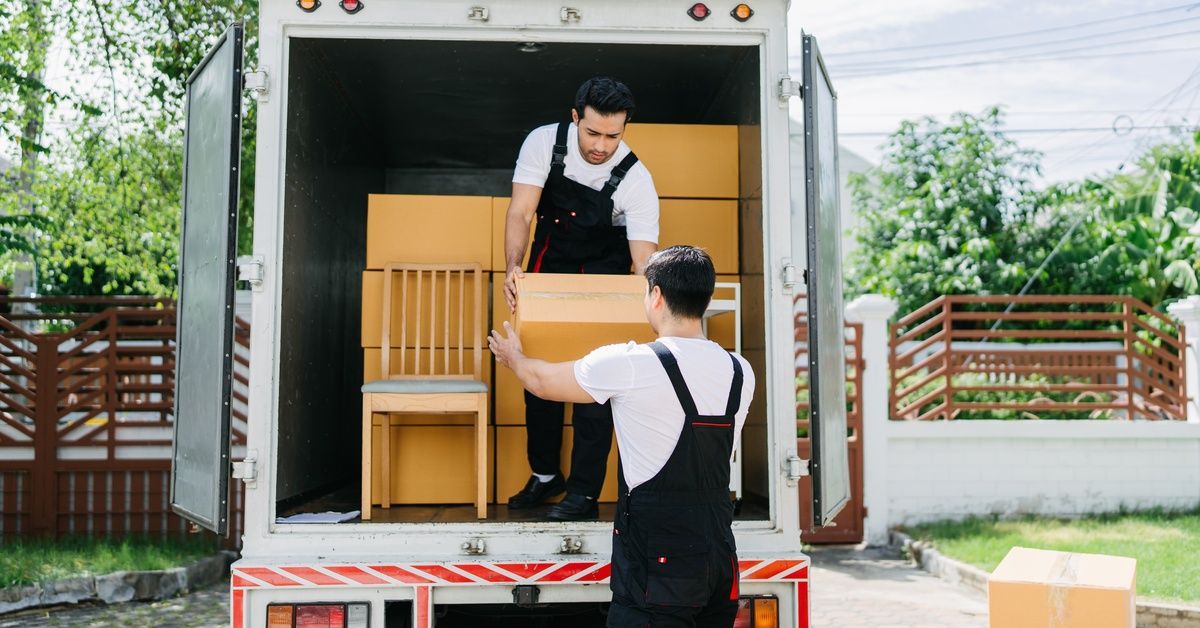 Two men in overalls are working together to unload a truck full of cardboard boxes. There is also a 