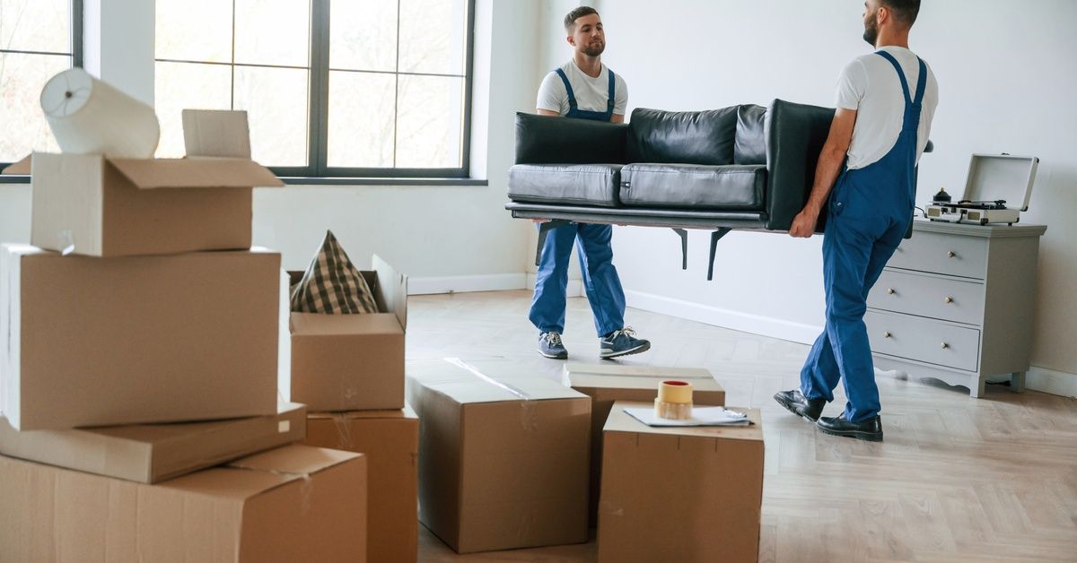 Two men in overalls are both holding a couch, moving it out of a room. Both men have short hair.