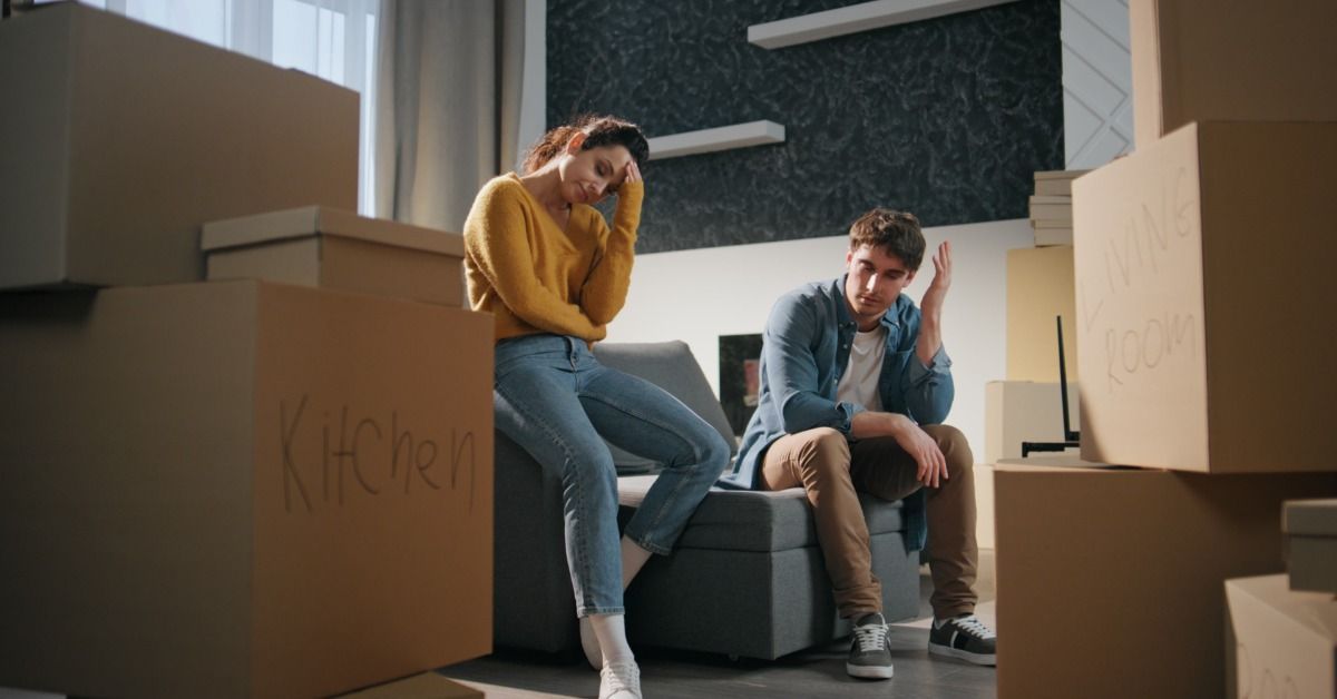 A man and a woman are sitting among piles of cardboard boxes, both of them looking distraught.