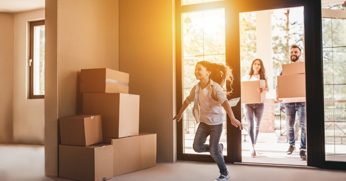 A man and a woman are carrying boxes while they and a small child walk into a home. The child looks excited.