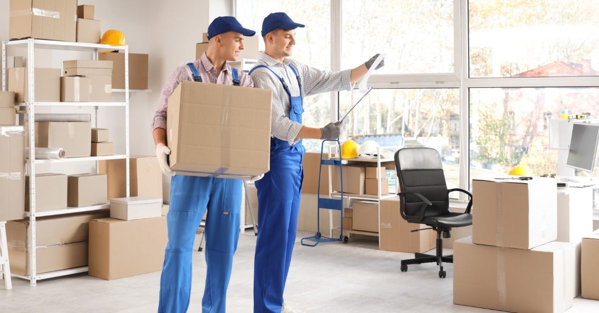 Two men, both of whom are wearing blue overalls, are standing in the middle of an office area.