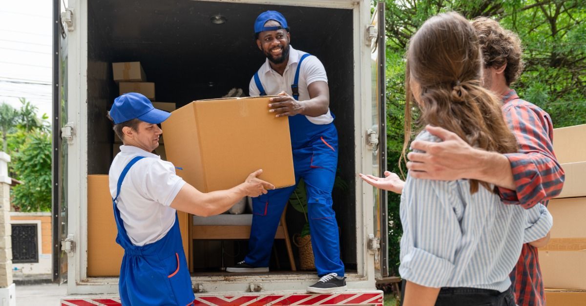 Two men are taking boxes out of the back of a truck. There is a man and a woman watching them work.