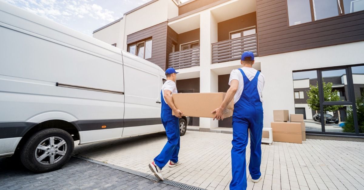 Two men, both of whom are wearing blue overalls, are carrying a box together into a home.