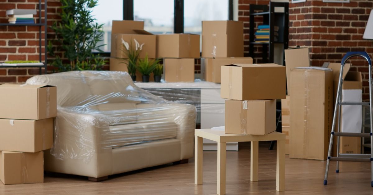 A living space has a number of moving boxes strewn throughout the room. There is a couch that is wrapped up.