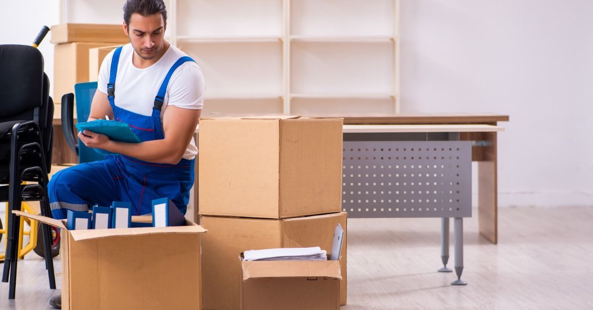 A man in blue overalls is kneeling next to several cardboard boxes. There are shelves in the background.