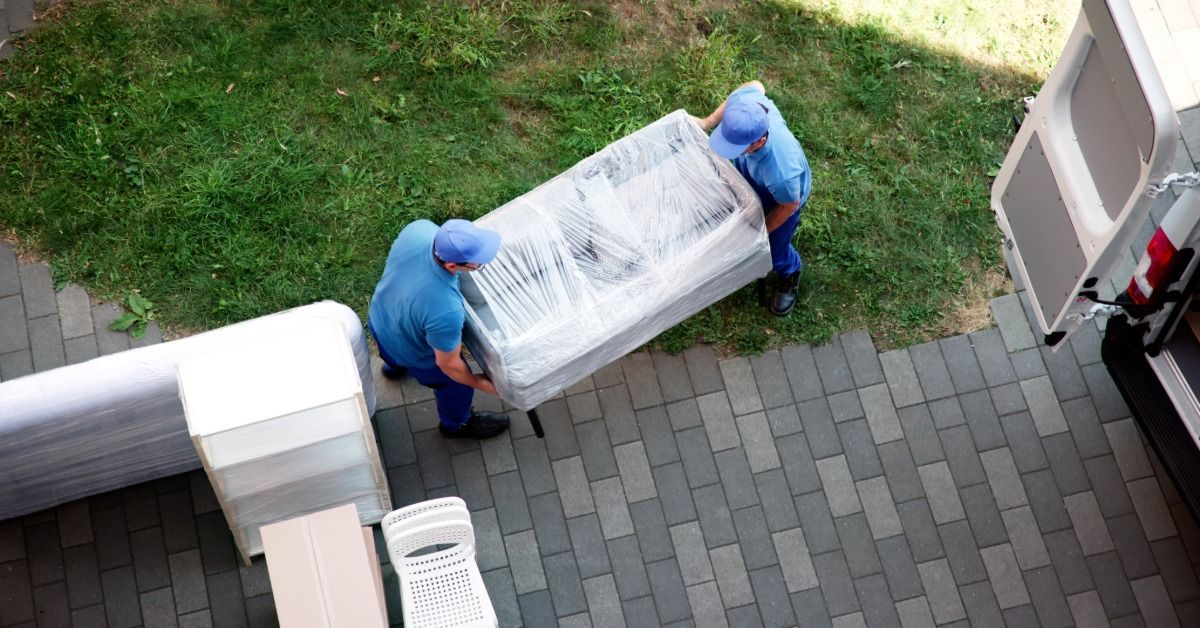 Two men, both of whom are dressed in pale blue, are lifting a couch together.