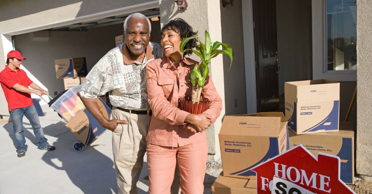 An older couple is standing in front of a house with a for sale sign. There are movers in the background.
