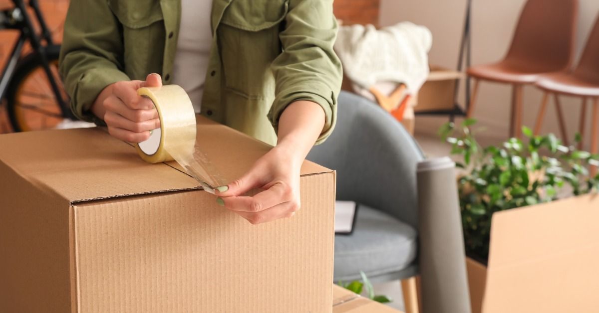A woman is using a roll of tape to tape up a cardboard box. She’s sitting in a living room.