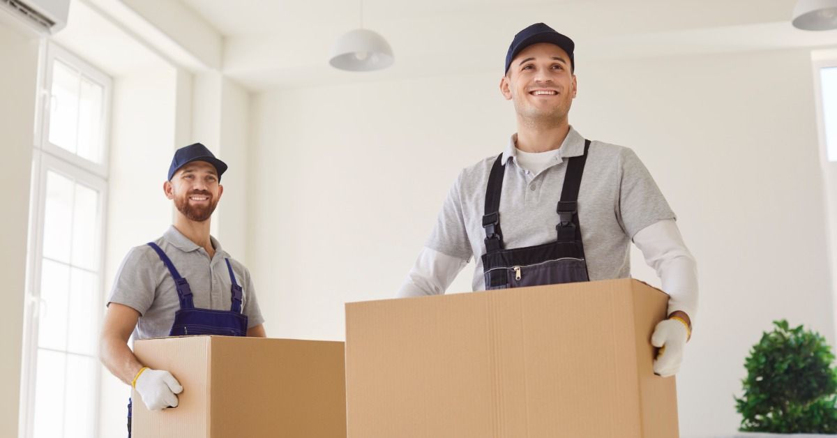 Two men are standing in a living room, and each of the are holding boxes in their hands.