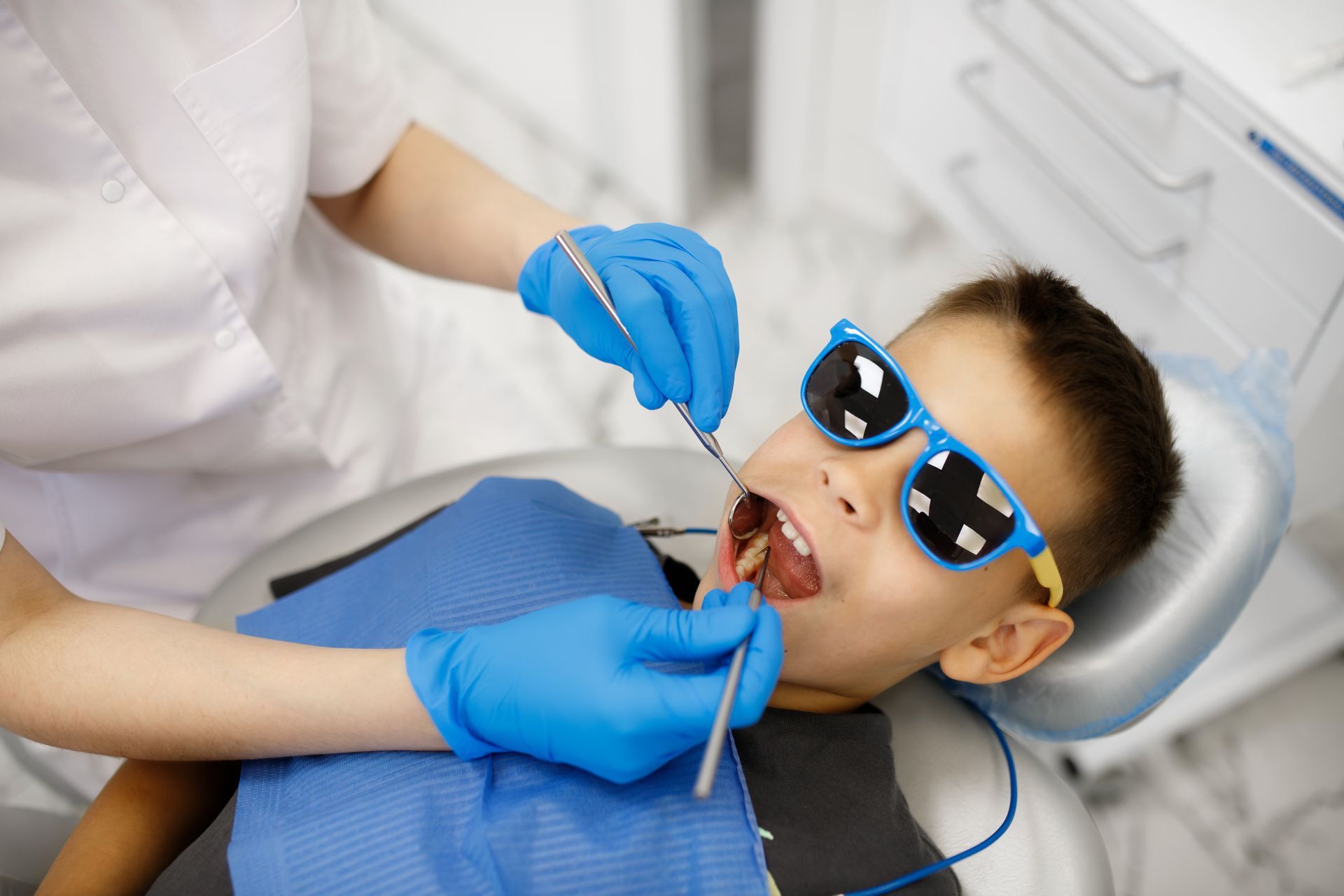 Young boy wearing sunglasses opens wide while a pediatric dentist examines his teeth with a mirror