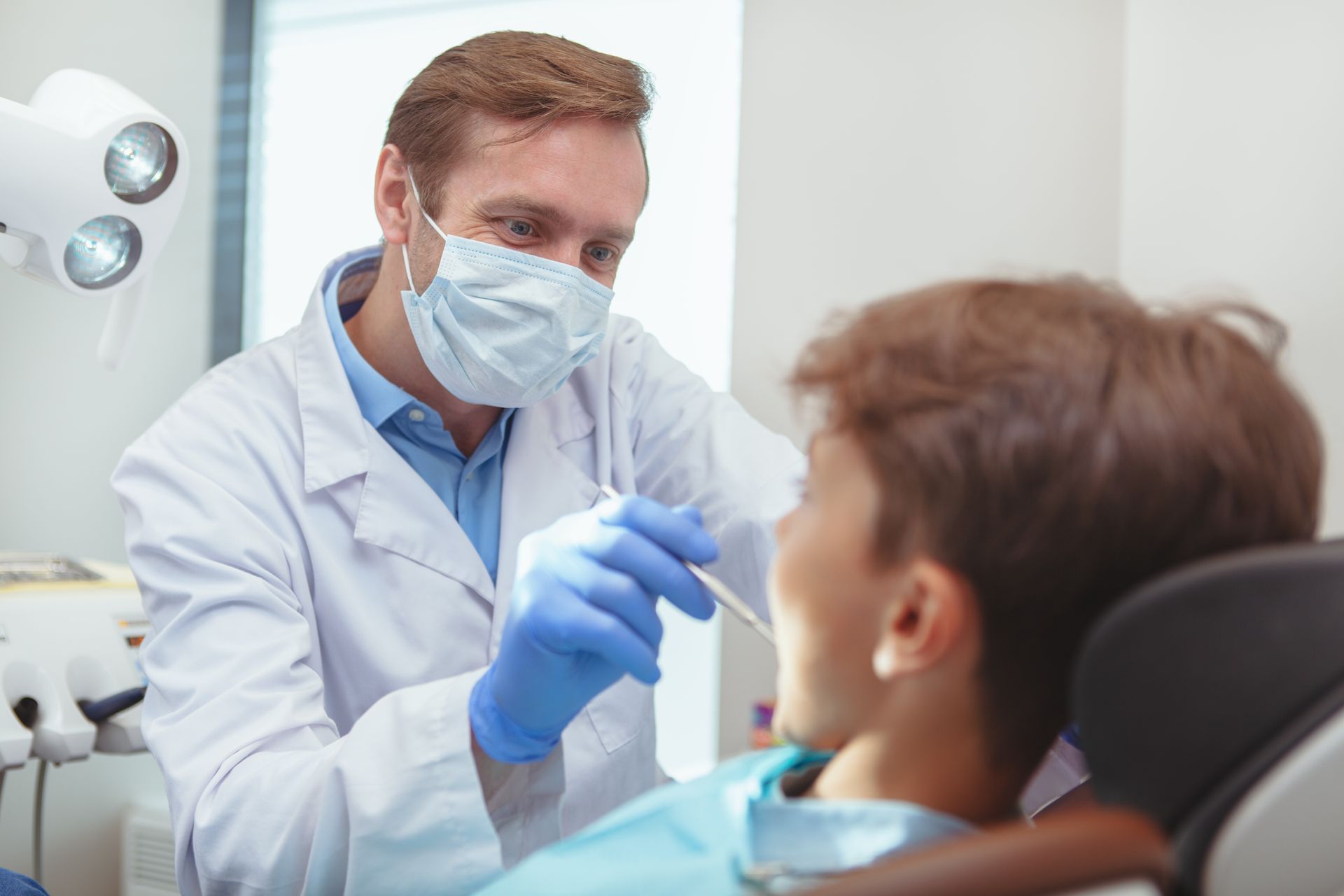 A mature male dentist wearing a medical mask and gloves, examining the teeth of a little boy.