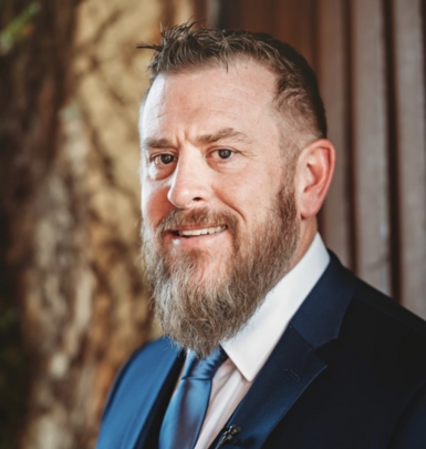 Man with a full beard wearing a blue suit and tie, smiling, indoors.