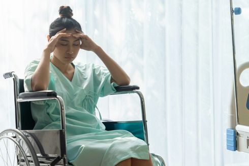 Woman in a mint hospital gown sits in a wheelchair, holding her head in distress by a bright window