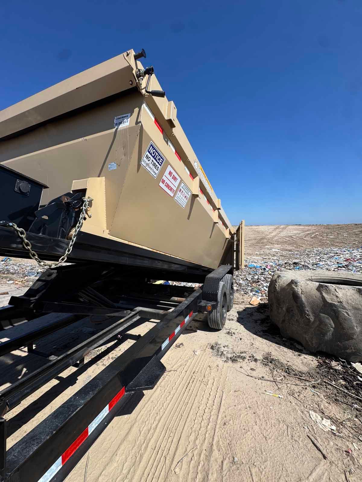 A tan dump container being lowered from a black trailer at a landfill under a clear blue sky.