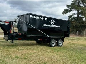 A black Diamond M Services dump trailer hitched to a truck in a grassy field under a cloudy sky.