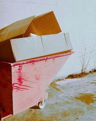 A pink dumpster holding cardboard boxes sits on a concrete surface against a white wall.