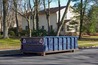 A large, empty blue dumpster sits on a residential street in front of a beige two-story house surrounded by bare trees.