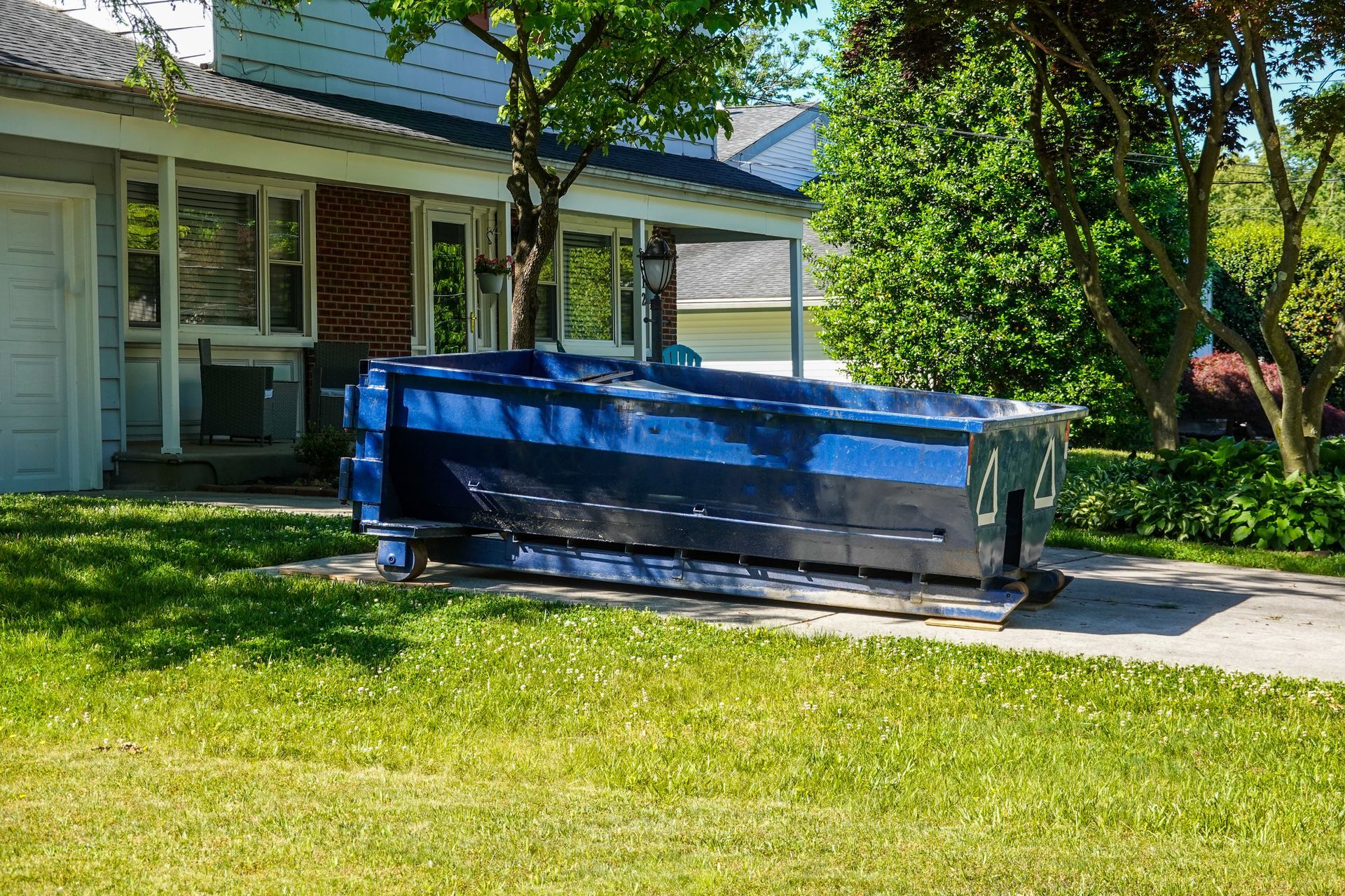 A large, blue metal dumpster sits on a concrete driveway in front of a residential house on a sunny day.