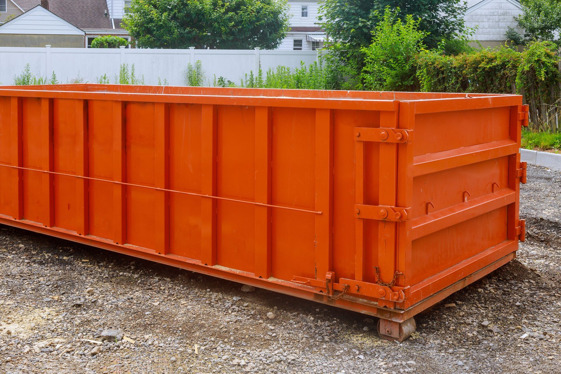 A bright orange, rectangular dumpster sitting on a gravel surface outdoors with a white fence in the background.