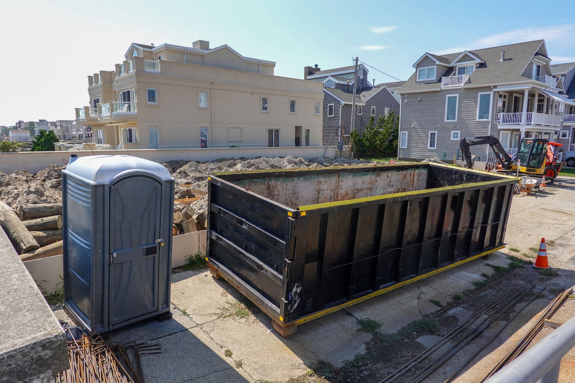 A portable toilet and a large black dumpster sit on a construction site next to residential homes.