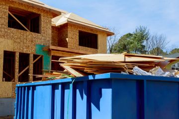 A blue construction dumpster filled with wooden scrap sits in front of a house under construction with exposed framing.