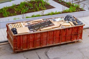 A large, rectangular red dumpster filled with construction debris and wooden scraps, positioned on a paved street.