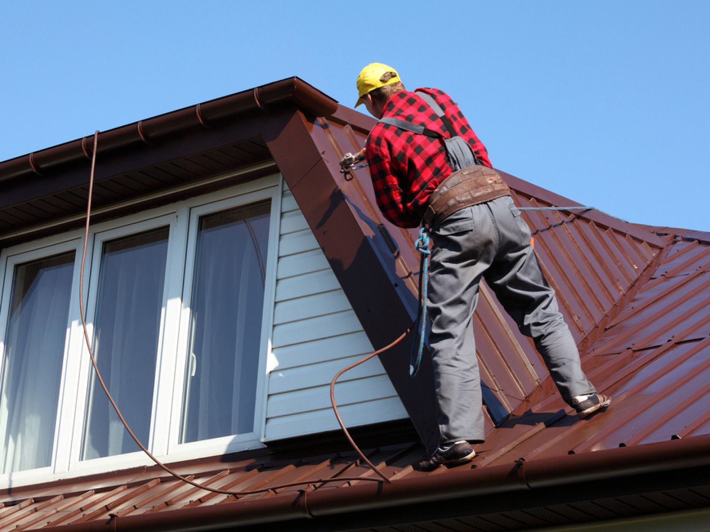 Worker on a red roof using a ladder near a white dormer window under a blue sky