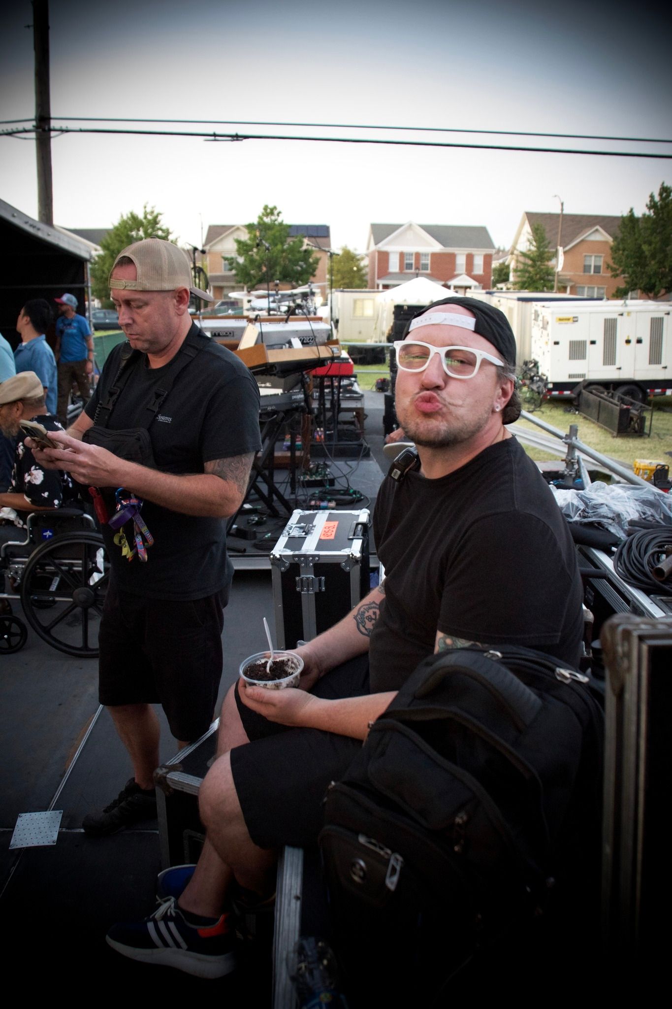 Two men at an outdoor event. One pouting, wearing glasses and a hat; the other using a phone.