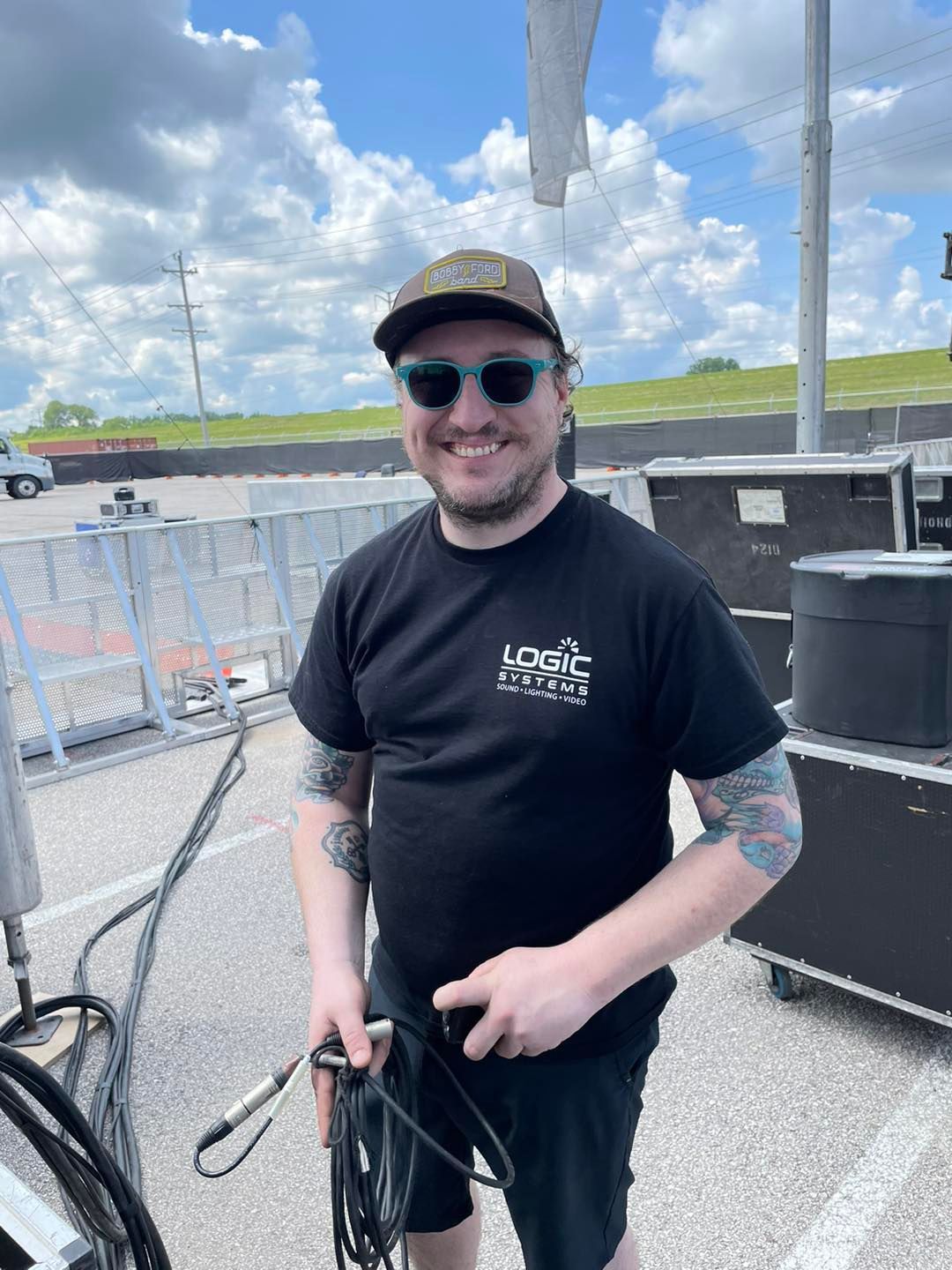 Man in black t-shirt, hat, and sunglasses smiles outdoors. Holding a cable.