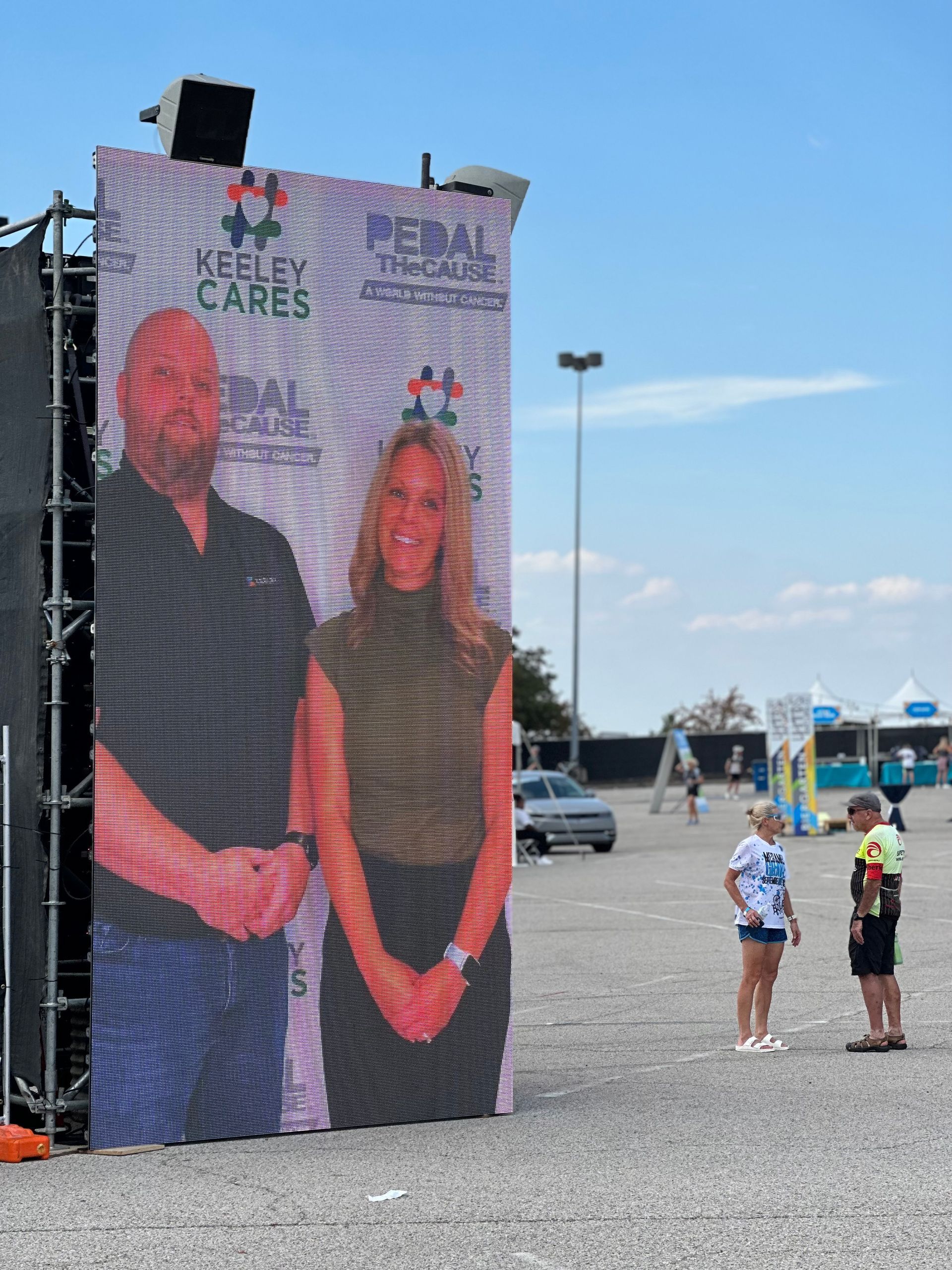 Large outdoor LED screen displaying a man and woman with text, two people watching, outdoor setting.