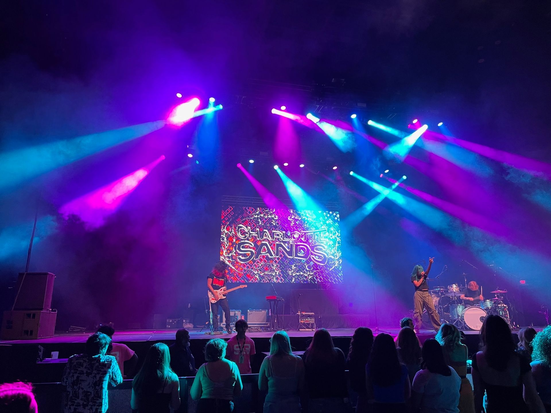 Concert stage with band lit by blue and pink lights, performing before a crowd.